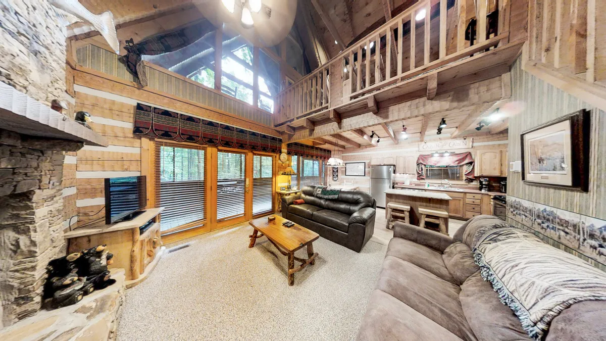 An inviting living room with a cozy stone fireplace, large windows offering picturesque forest views, comfortable sofas, and access to a wooden balcony. The ambiance is warm and romantic, ideal for couples seeking comfort after a day of adventure.