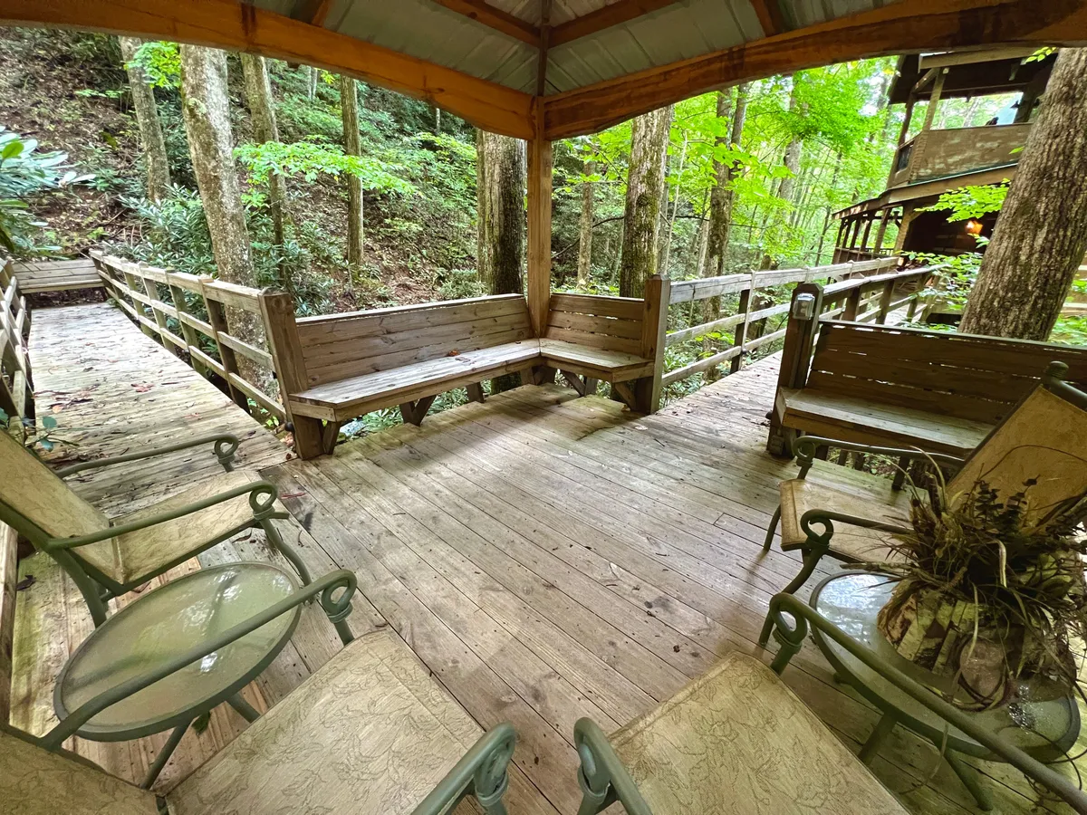 This image showcases an inviting exterior deck area with rustic wooden seating and railings set against a lush backdrop of trees. The seating is oriented to provide tranquil views of the natural surroundings, perfect for relaxing conversations or simply enjoying the peaceful ambiance of the woods.