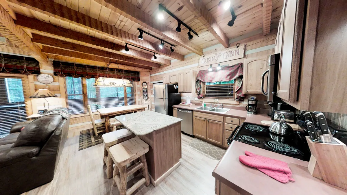 A spacious, welcoming kitchen and dining area featuring prominent wooden beams, a large kitchen island, big windows letting in natural light, and classic cabin decor. The open layout makes it perfect for hosting group meals, cooking together, and socializing.