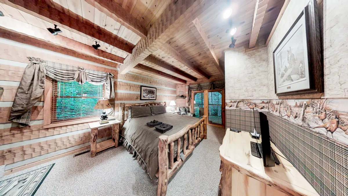 A cozy bedroom with a rustic log bed, exposed wooden beams, a window that frames a nature view, and carefully chosen decorative touches. Bedside lamps and wall art contribute to the inviting, restful mood.
