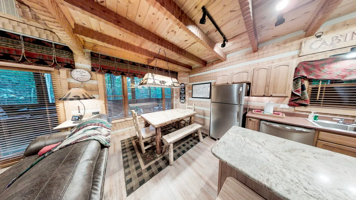 A view of the open-concept kitchen, dining area, and living space. A large wooden dining table is perfect for family meals or game nights, and the adjacent kitchen features modern appliances and ample workspace. Leather sofas provide cozy seating nearby, while overhead wooden beams and windows tie the space to its natural surroundings.