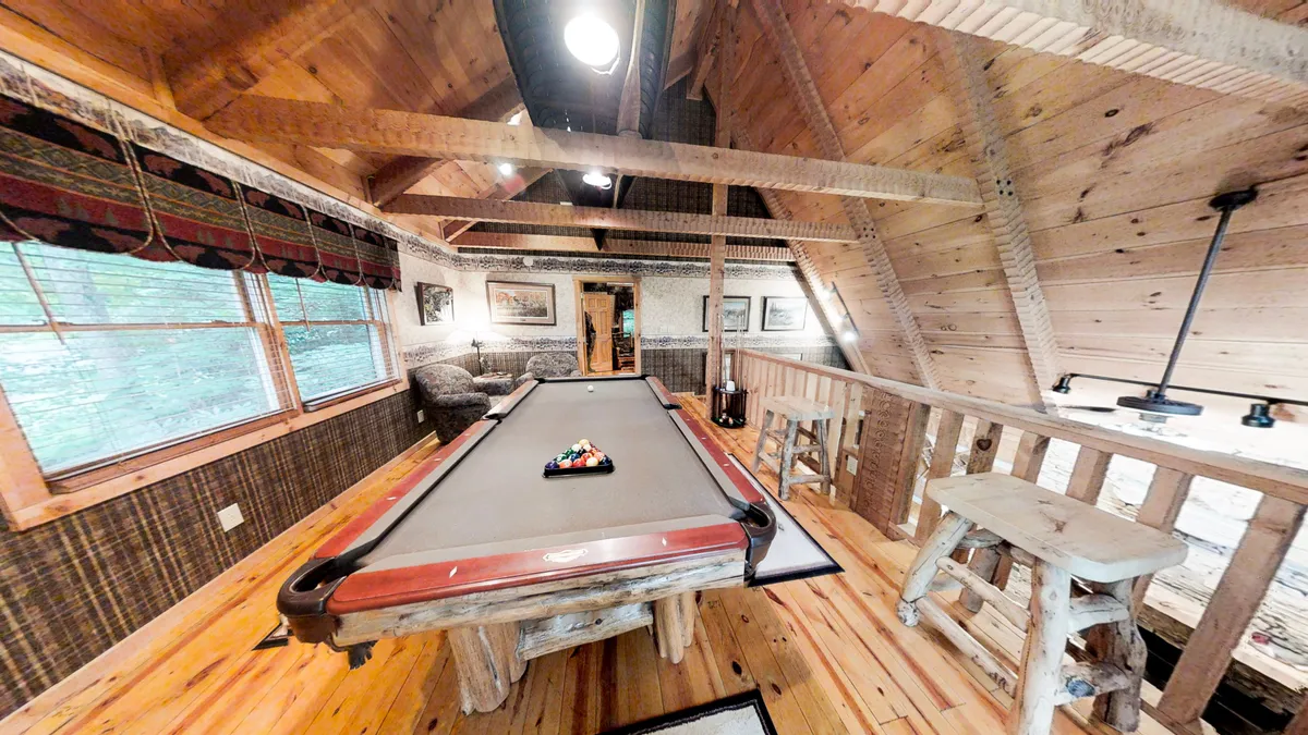 A warmly lit game area with a prominent pool table, exposed wooden beams, and a nearby comfortable seating nook. Framed artwork and windows add a homely touch, making this an ideal spot for both competition and relaxation.
