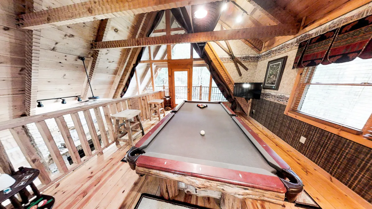 A vibrant loft game room featuring a full-size pool table taking center stage, surrounded by large windows that bathe the space in natural light. Rustic wooden beams overhead and comfortable seating create an inviting, playful atmosphere perfect for gatherings.
