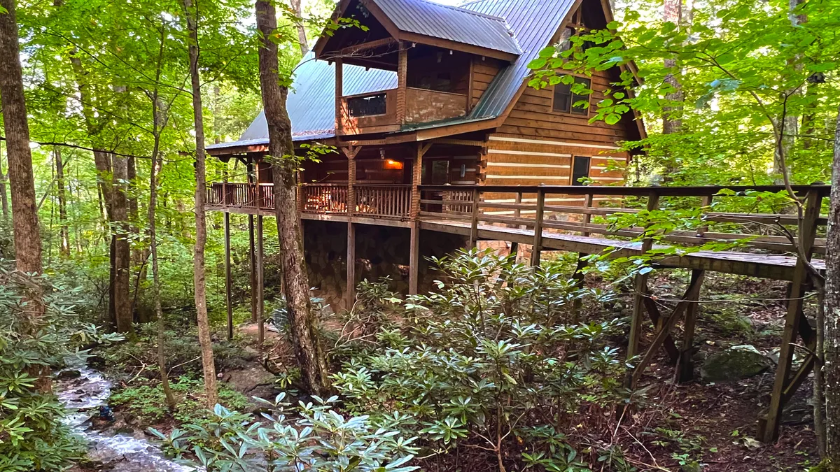 The hero image features a charming log cabin nestled among lush trees in a serene woodland setting. A wooden walkway curves invitingly toward the front porch, while a gentle stream meanders nearby, creating a peaceful, secluded vibe. The natural beauty of the forest surrounds the cabin, hinting at abundant opportunities for relaxation, wildlife watching, and outdoor adventures.
