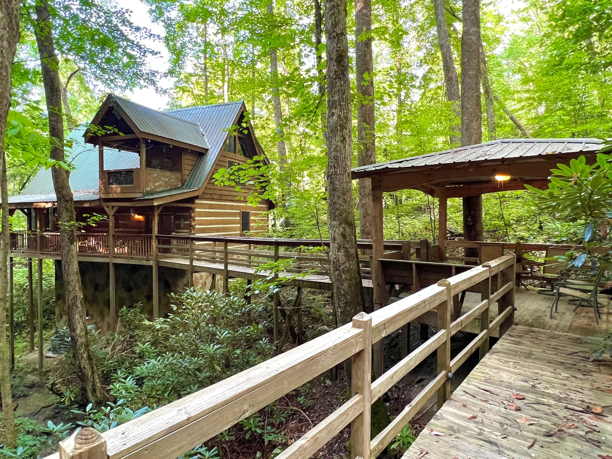 The image displays a rustic log cabin nestled amongst lush greenery, featuring a spacious wooden deck and an inviting open gazebo. The natural surroundings create a sense of seclusion and relaxation, while the deck and gazebo suggest spaces for family gatherings, outdoor dining, and stargazing after a day of adventure.