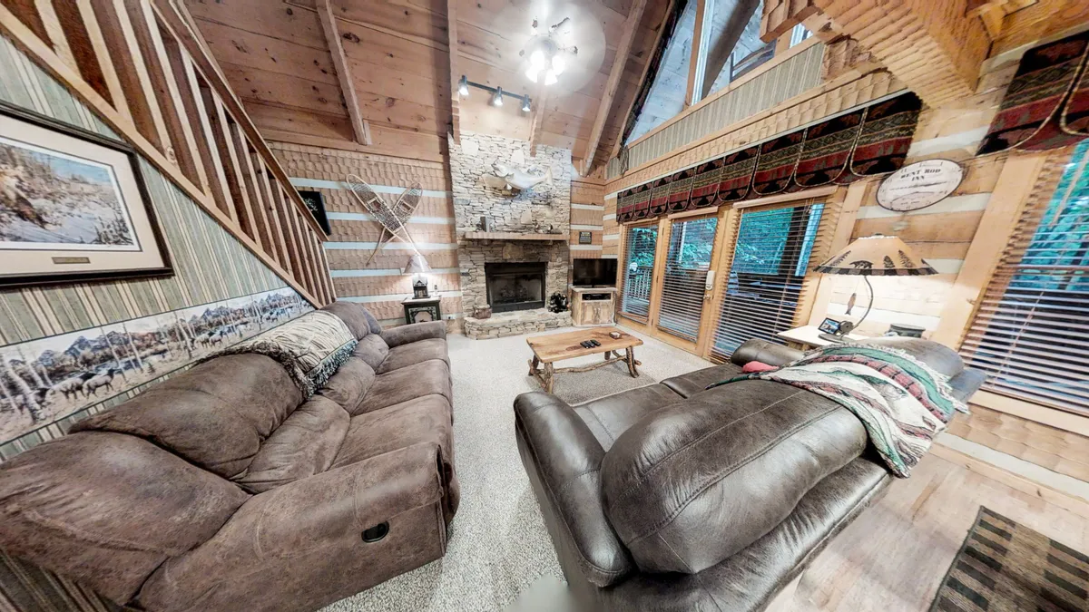 A spacious, open living room featuring a striking stone gas fireplace surrounded by comfortable sofas. Large windows allow natural light to flood the space and offer tranquil creekside and forest views. Exposed wooden beams add rustic charm, creating an inviting, communal hub for relaxation and connection.